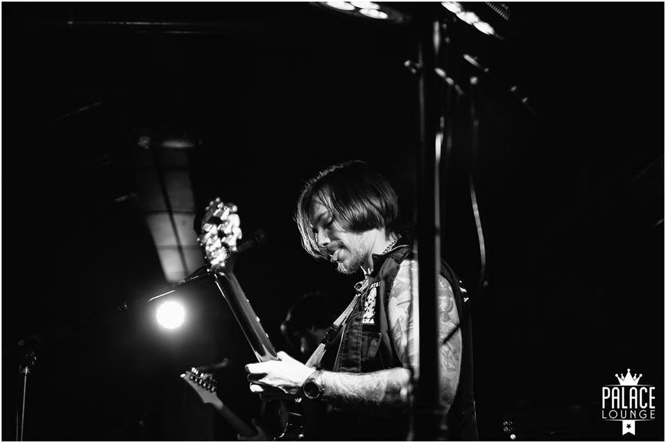 Reggie leans back on stage with his electric guitar, his face tense with concentration as he solos. His hair is in his face and his tattooed arm is visible through his patchwork best