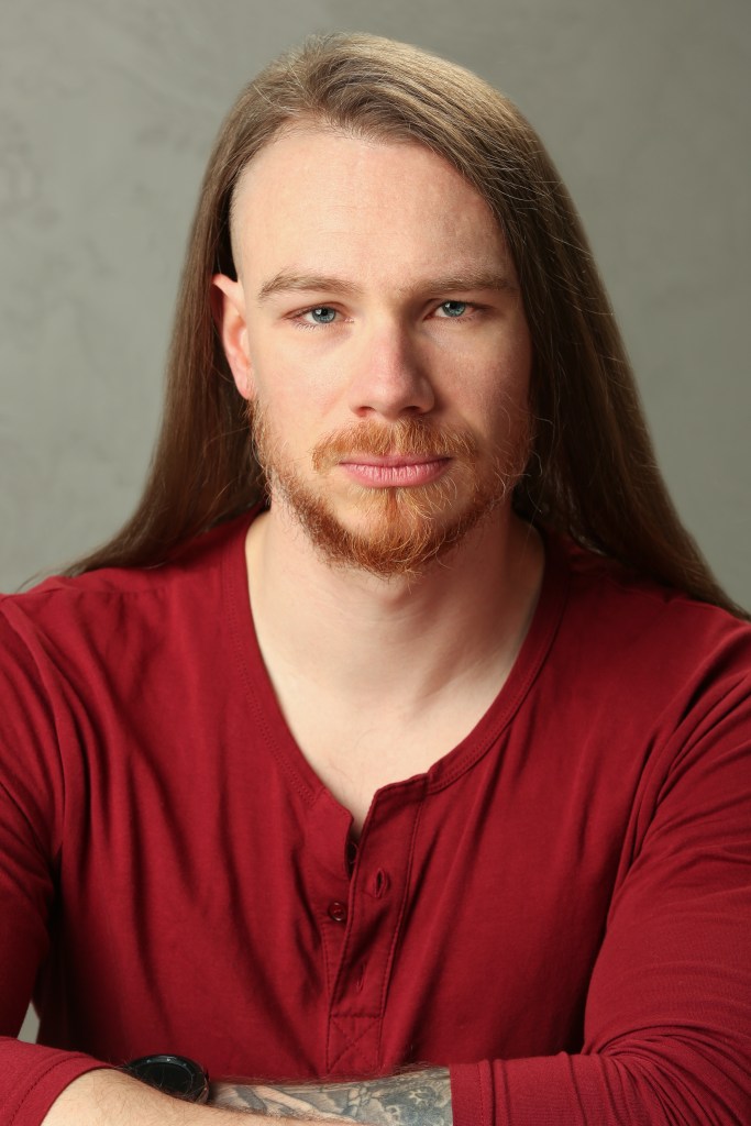Headshot of Reggie Herbert, a white male in his early thirties. He has long brown hair undercut on the sides, striking blue eyes, a reddish brown trimmed beard and mustache. He gazes intently into the camera, his strong jaw set.